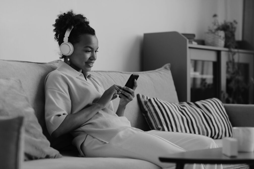 A woman sitting on a couch, wearing headphones, and smiling while listening to a podcast on her phone.