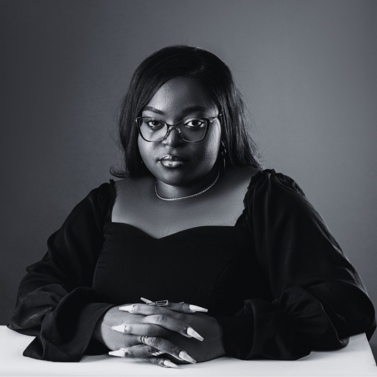 Deborah Alifa, Lustnfree counselor, seated with folded hands at a table, wearing a dark long-sleeve top and necklace, in front of a neutral gray background.